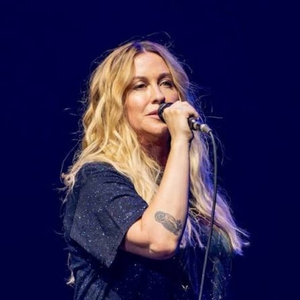 Alanis Morissette singing into a handheld microphone against a dark blue stage background.