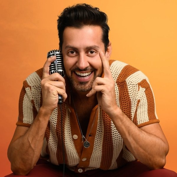 Max Amini smiling and holding a microphone beside his face against an orange background.