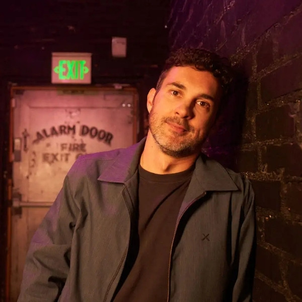 Mark Normand leaning against a brick wall beside a backstage exit door.