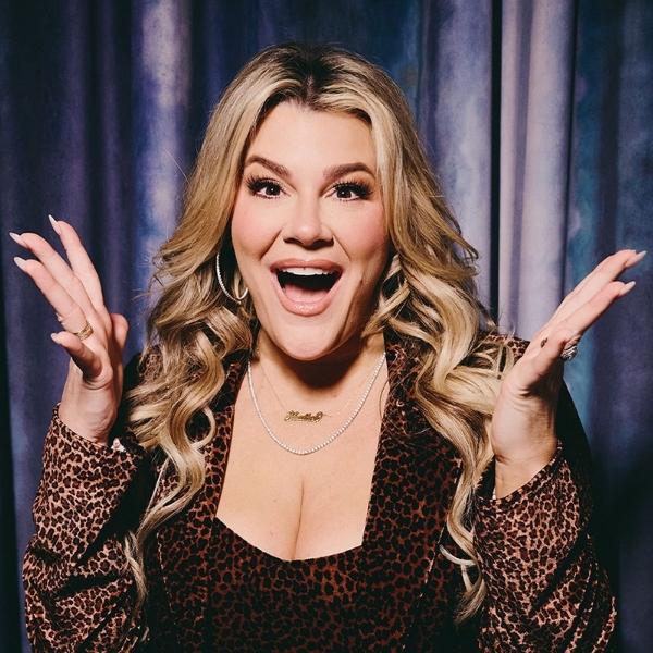 Heather McMahan smiling with both hands raised, wearing a leopard-print outfit in front of dark curtains.