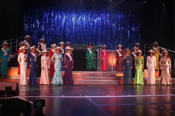 Rodeo pageant contestants wearing cowboy hats, sashes and evening gowns stand onstage beneath blue lights.